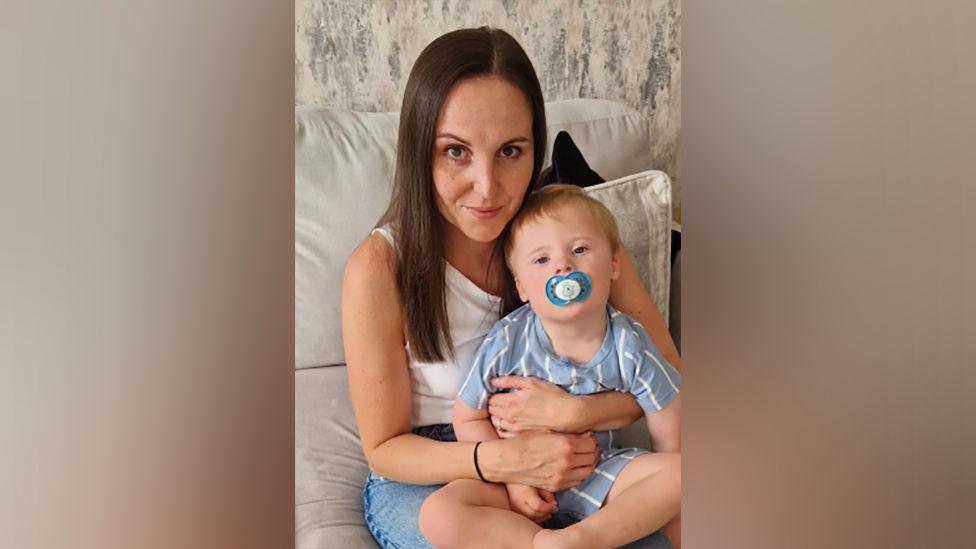 Kirsty Pedelty, with long brown hair, holds her son, who is blonde with a dummy in his mouth and wearing a striped suit