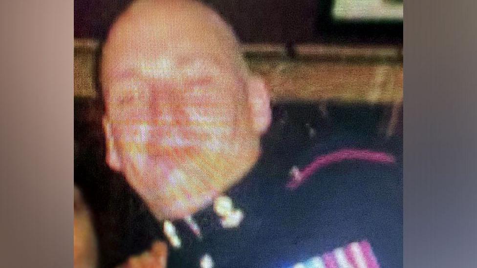 A blurred image of Michael Webber - he is wearing a dark-coloured uniform and has medals on the left side with ribbons of purple/white and blue/white. He is bald.