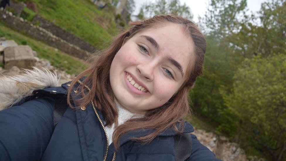 Christie Harnett is taking a selfie and smiling broadly at the camera. She has long brown hair and is wearing a blue coat.