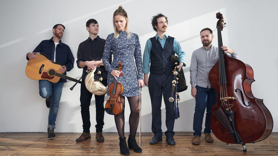 Five members of the band Breabach standing on a wooden floor with their instruments. A woman in the foreground carries a fiddle while two men either side of her have bagpipes. Another man has an acoustic guitar, another a double bass. 