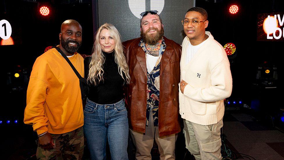 Four people - a man, a woman and two men, stand in a line with their arms around each other. They are in a darkened studio environment. The man in the centre of the group wears a long, brown/red leather jacket over a shirt with a colourful pattern. His chest is heavily tattooed and he also has tattoos on his face.