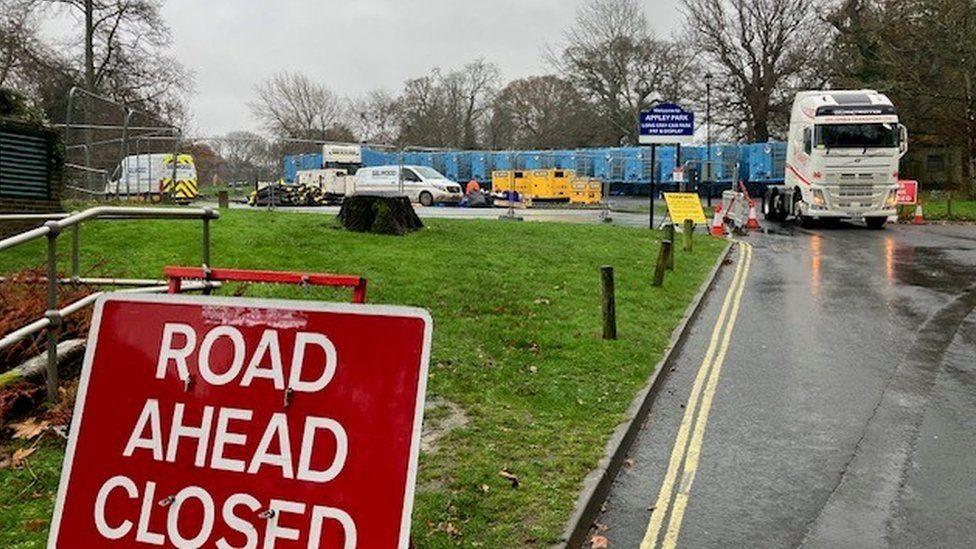 Road closure sign, white lorry cab at entrance of car park with long line of blue containers set up.