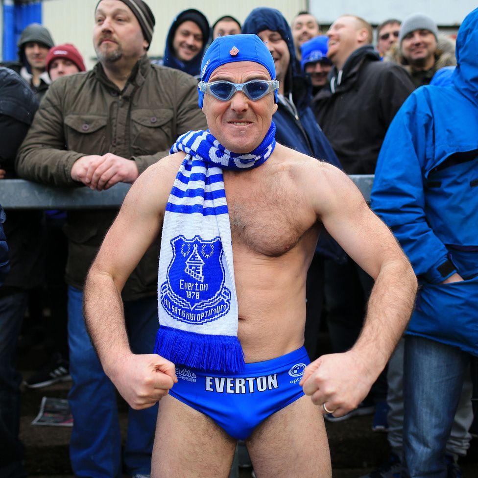 Speedo Mick doing a bodybuilder pose in his trunks, an Everton scarf, swimming cap and goggles, on the terraces surrounded by fully clothed fellow fans at Everton's FA Cup game at Carlisle United in 2016. 