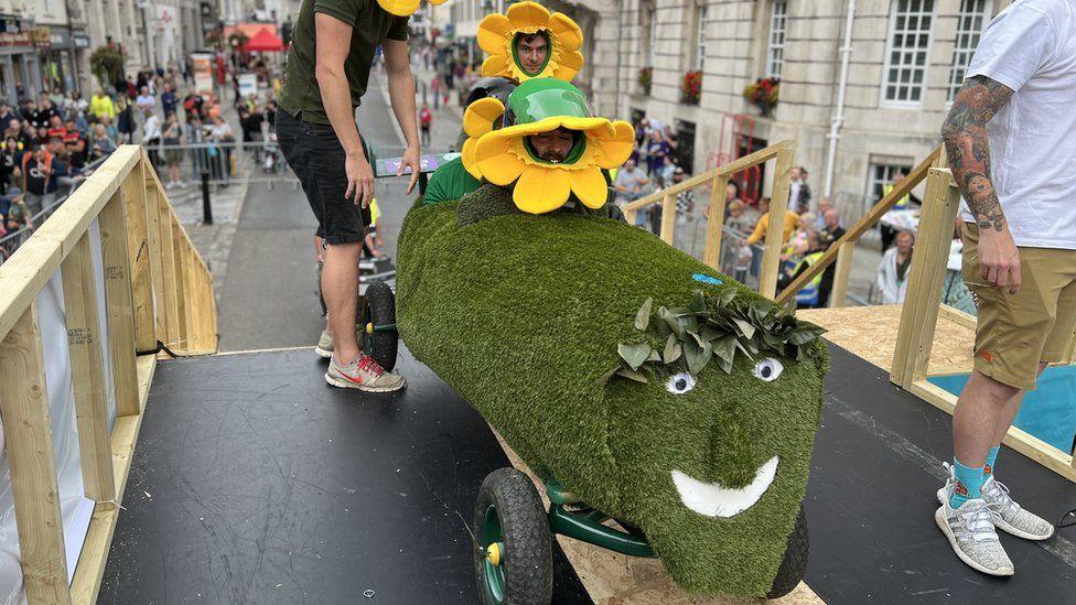 Two men sitting on their kart, which is made of astro turf and designed to look like grass. The men have yellow sunflowers on their heads and they are at the top of a ramp.