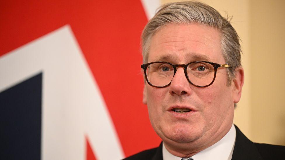 Sir Keir Starmer, with grey hair and dark glasses, sits in front of a union flag in a close-up shot. 