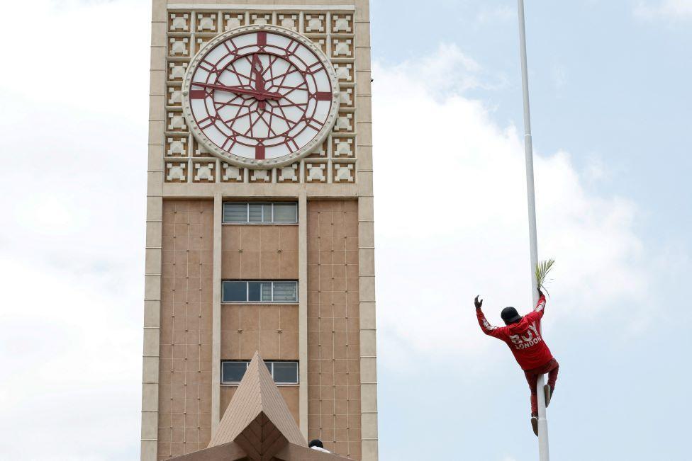 A man climbs up a tall pole with the parliament tower seen behind it.