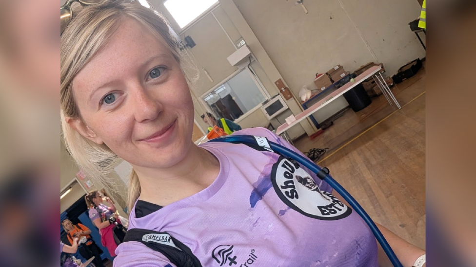 Enlli Williams has blonde hair with a side parting tied in a ponytail and is looking at the camera in a selfie, smiling. She has sunglasses on top of her head. She wears a purple running top with the SheUltra logo partly visible. Straps from a bag are going over her shoulder. She is standing in a hall with some tables and boxes present. A few competitors and people in high-vis jackets are in the background.