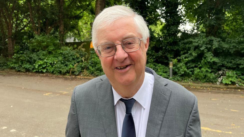 Mark Drakeford, the cabinet secretary for finance and Welsh language in the Labour Welsh government, smiles into the camera, wearing a grey suit and white shirt. There are a row of trees in the background.