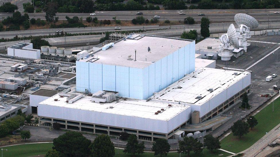 A rectangular blue building surrounded by outbuildings and roads. Two large satellite dishes can also be seen.