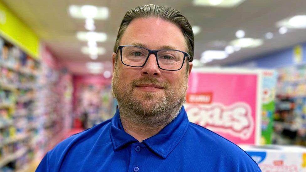 A head and shoulders shot of Lee James Adlam looking straight at the camera.
He wears the royal blue shirt which the uniform of The Entertainer and glasses. He has a beard and moustache. Behind him are the blurred outlines of boxes of toys.