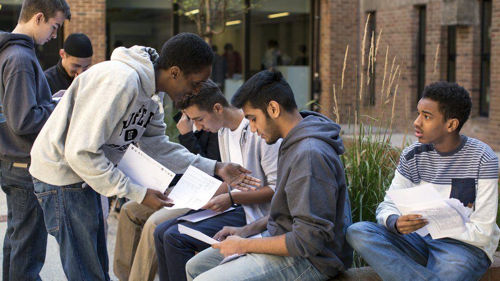 A group of male students sit outside a school or college building opening their exam results.