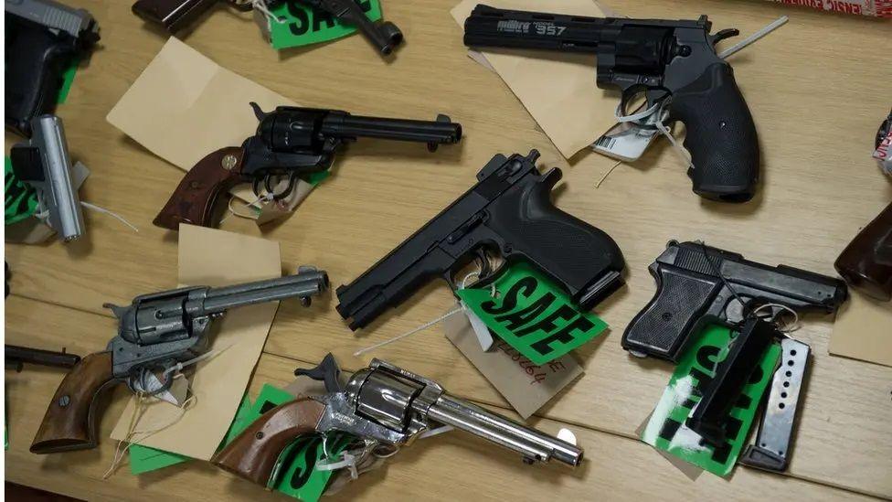 Various pistols on a table. Some are black and one is grey. Some have green labels attached which say "SAFE". Some have light brown slips of paper attached.