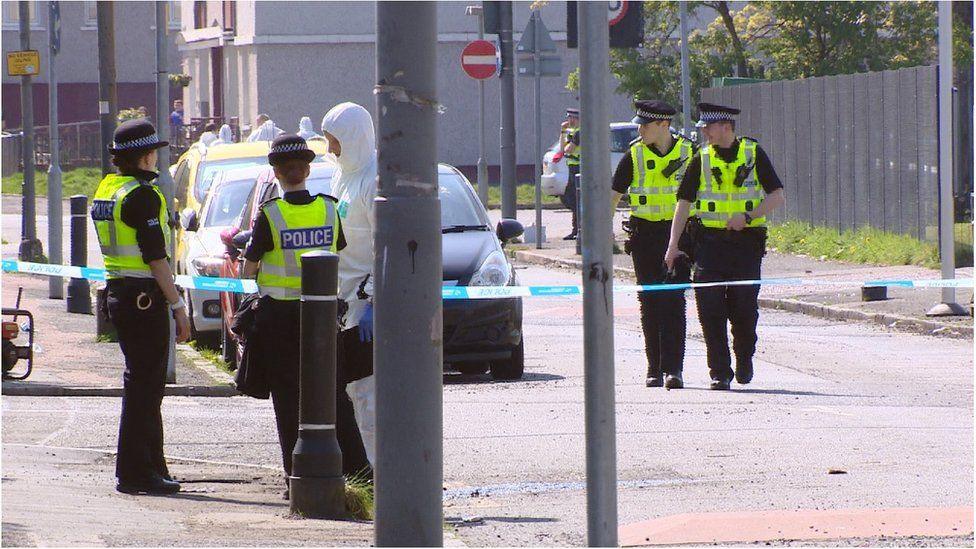 Police and forensic officers walk around a quiet street behind a cordon. Cars are parked on one side of the road 