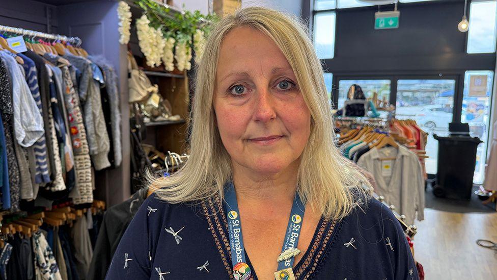 A woman with blonde shoulder-length hair. She is wearing a St Barnabas lanyard and a dark blue dress with a white dragonflies pattern.