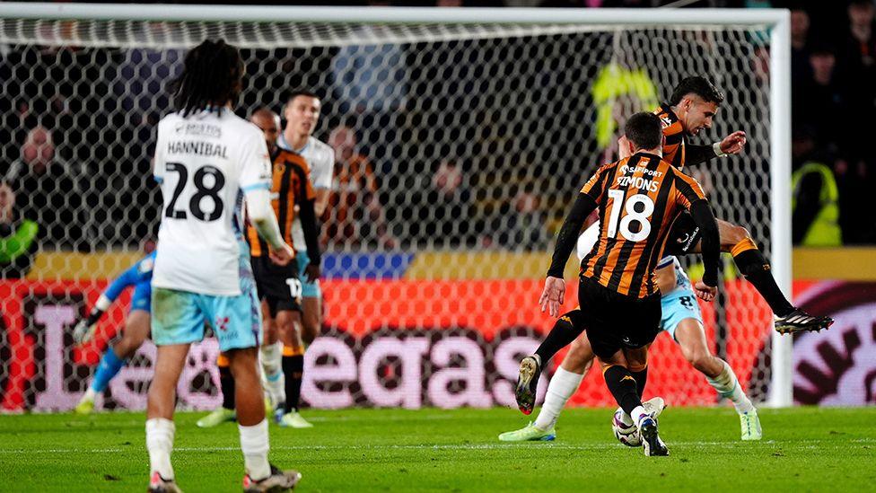 Hull City Xavier Simons in black and amber shirt and black shorts surrounded by other players scores the opening goal against Burnley 