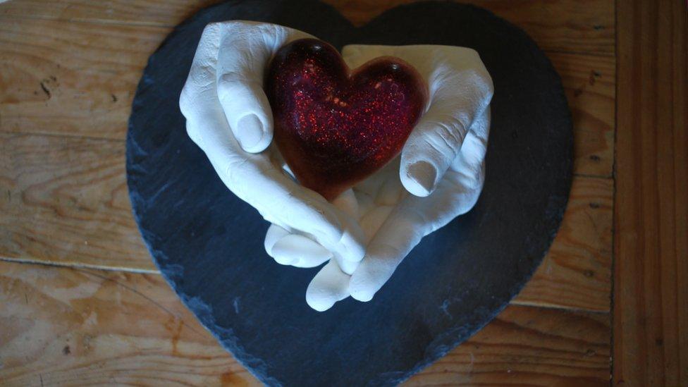 A cast of Kelly and Paul's hands, holding a red heart made from breast milk, resting on a heart shaped slate