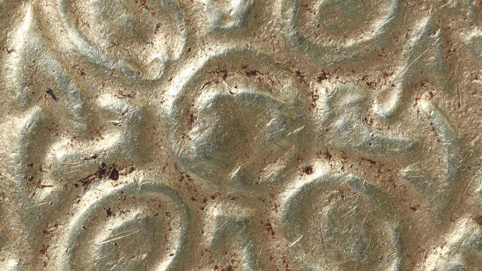 A very tight close up of the front of an early 8th Century gold round pendant. It shows the middle of the pendant with its slightly raised circular boss in the middle and the oval patterns radiating out from the middle.