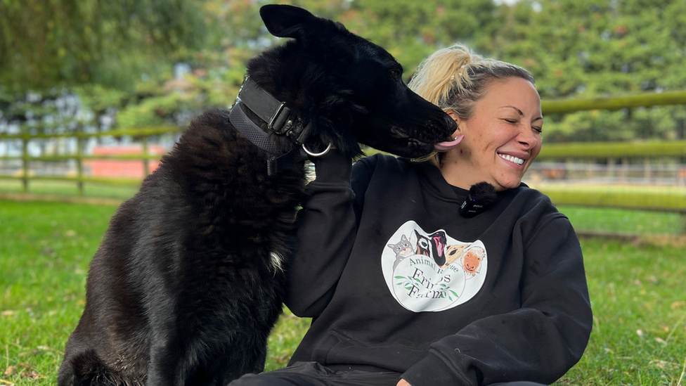 Jodie Marsh wears a black hoodie scrunches up her face and smiles as a black dog licks her ear.
