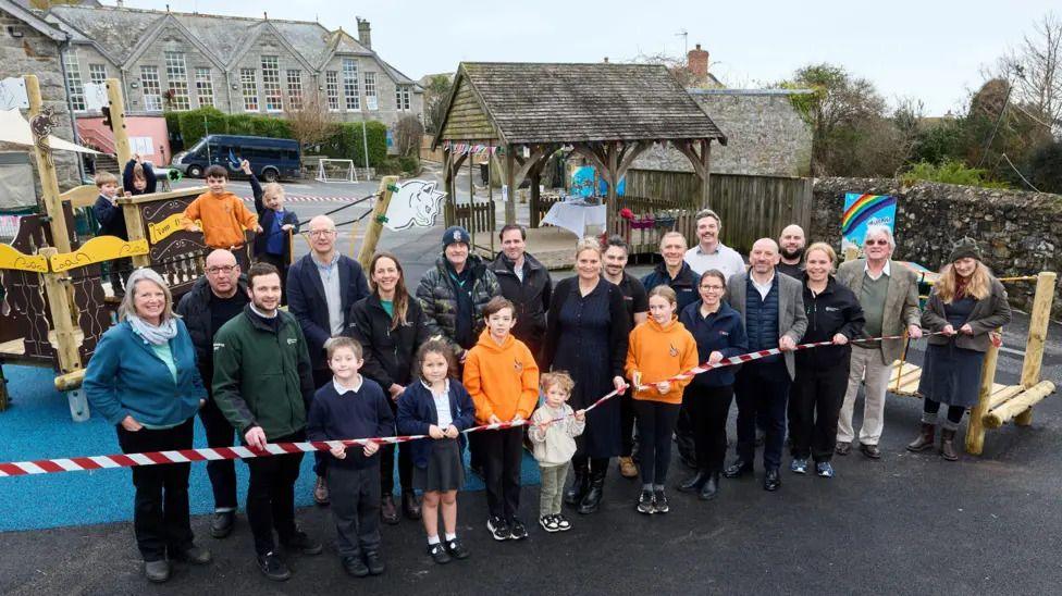 kids and teachers at the school standing behind a red and white ribbon and in front of the new playground.