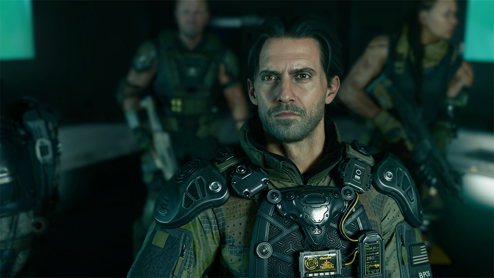 A screenshot shows a realistic rendering of actor Milo Ventimiglia's character wearing an elaborate military uniform and body armour. He's looking up, his face illuminated by the glow of a screen, as his squadmates look on in the background.