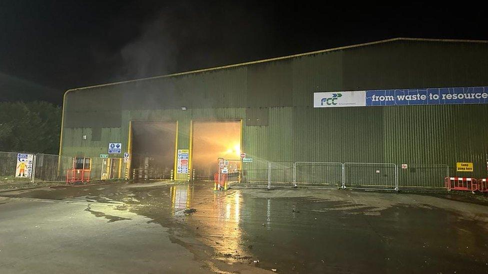 A corrugated metal building with open doors showing the bright orange fire blazing inside.