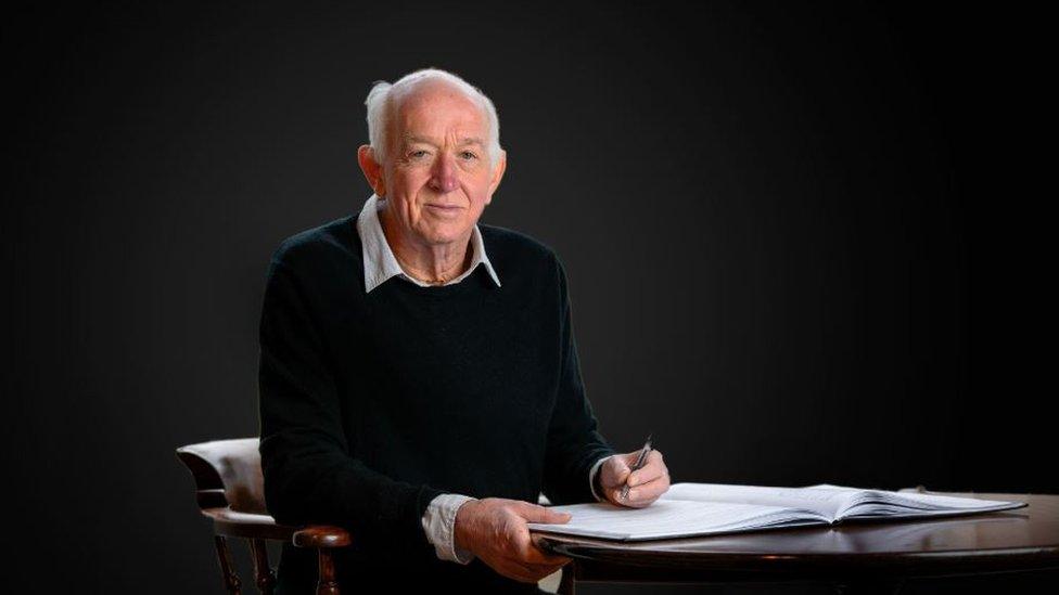 A man with white hair sitting on a chair in front of music sheets