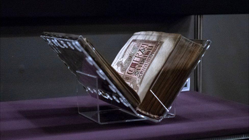 The Lindisfarne Gospels on display at the British Library. It is resting on its spine in a clear acrylic stand, open to show one of its pages on the right side. The page is illustrated with a colourful early medieval design and has Latin writing.