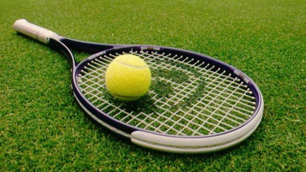 A tennis racket and a tennis ball laid on a grass tennis court.