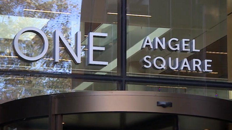 White lettering saying "One Angel Square" in glass windows above a brown-framed revolving door to a public building.