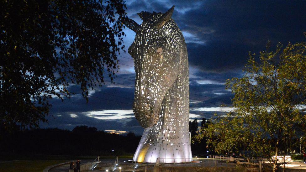 A large sculpture of the Kelpie in silver against a dark and cloudy sky. The sculpture is lit from within and shafts of white light can be seen below. Two trees are in the foreground.