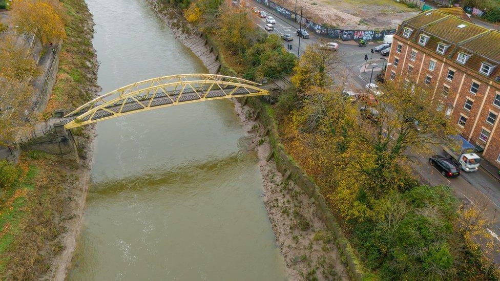 An aerial image of Langton Street Bridge. It is a yellow banana shaped footbridge across a river. There are roads on either side and trees lining the riverbanks, which are made from soft mud and silt. 