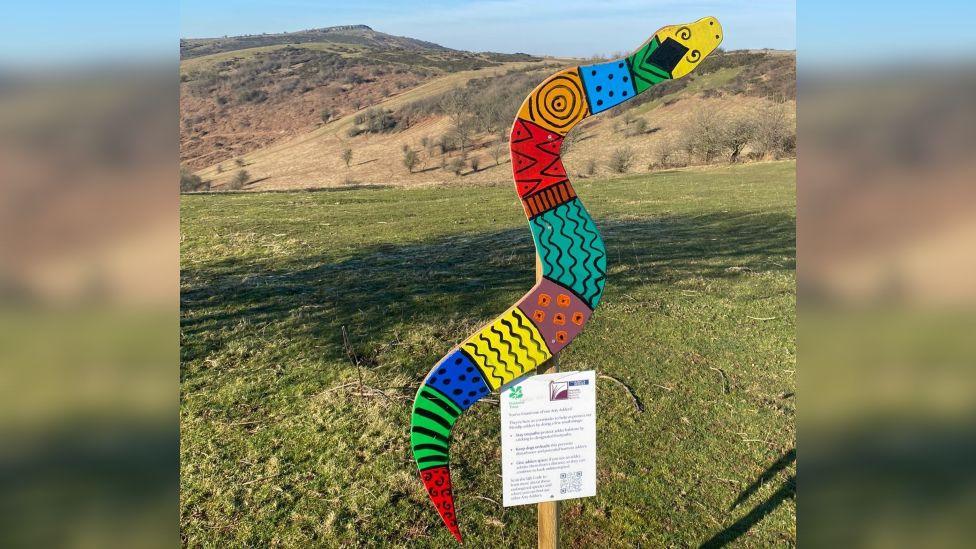 A large wooden snake painted in various bright colours and patterns. It is nailed to a wooden stick with an information sign and a QR code beneath it. In the background you can see the Mendip Hills on a sunny day.