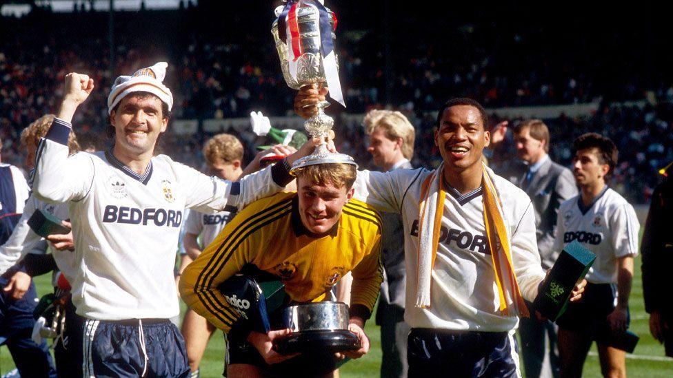 Luton Town captain Steve Foster in a white and blue strip with arms upraised; goalkeeper Andy Dibble in bright yellow strip and bending over, holding the trophy's base; and Brian Stein in the white and blue strip. Steve and Brian are holding the trophy up on Andy's head. They are grinning and standing on a pitch with other players and officials behind them and beyond them packed stands. 24 April 1988.