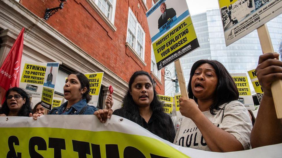 A row of striking nurses stand in their picket line at Guy's & St Thomas' Hospital, in London. Dressed in off-duty clothing, four women can be seen behind a yellow and white banner which spells out part of the hospital's name. One of the women is holding a yellow and black placard with the slogan "stop exploiting your nurses".