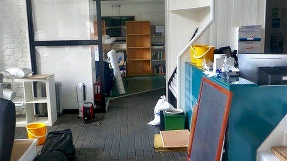 Furniture and fittings in storage including shelves, noticeboards, yellow buckets and books. The floor is clearly damp.