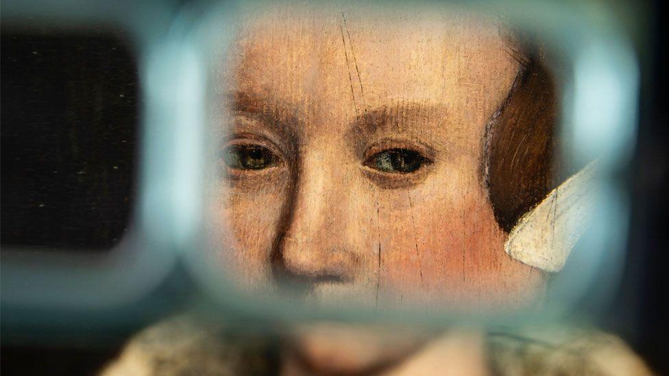 A close-up of a portrait of a Tudor woman believed to be Lady Jane Grey. She has reddish hair under a white linen cap. She is looking left. Her eyes and nose are framed and enlarged by a lens which shows lines which once slashed through the left eye.