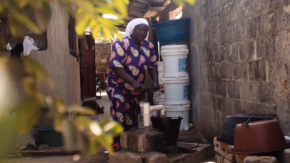 A woman next to a wall pumps water from a borehole in a bucket.