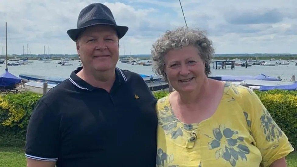 Stephen Baxter a man standing with his wife Carol Baxter. Mr Baxter is wearing a navy T-shirt and hat. Mrs Baxter has grey curly hair and is wearing a yellow top with grey flowers.