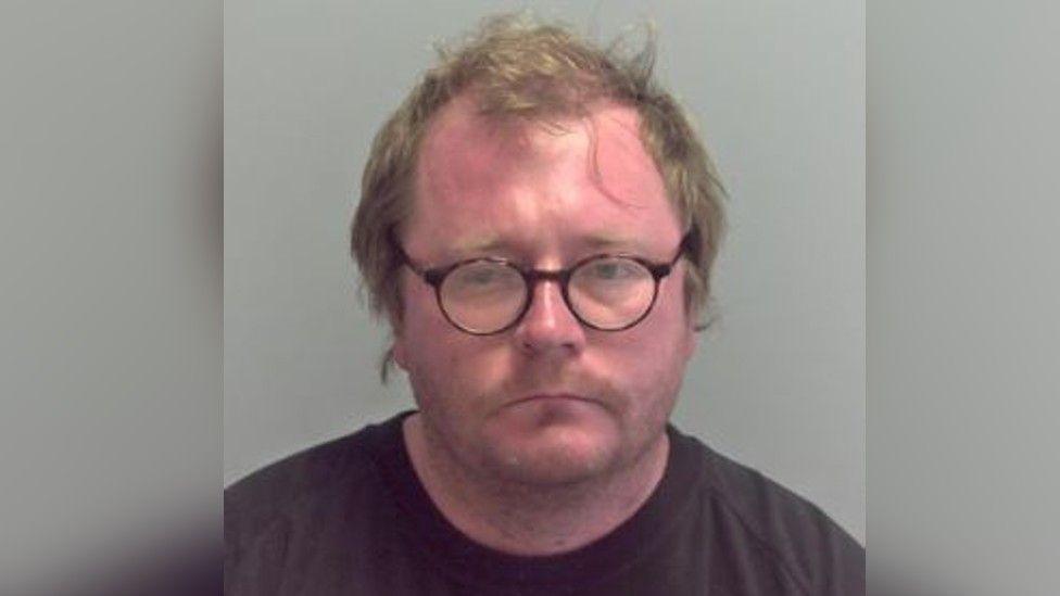 Allan Scott in his custody photo. He is wearing a brown round-neck T-shirt, has glasses, is unshaven, and his light brown hair is scruffy.