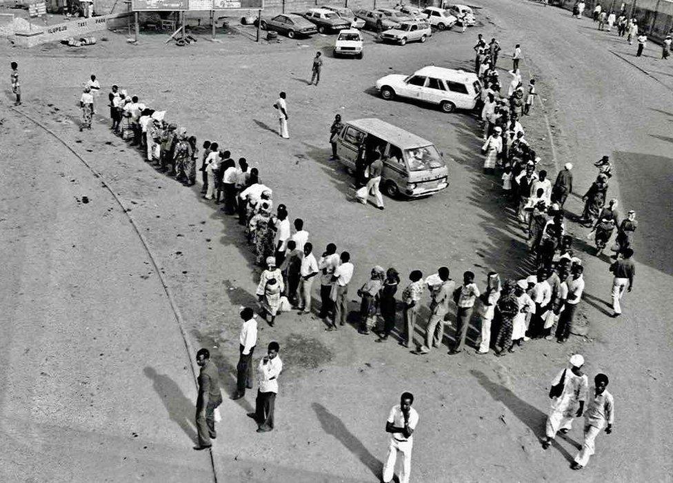 A photo by Sunmi Smart-Cole entitled: "Why... No Buses" - 1984, showing Nigerians waiting in a line for a bus