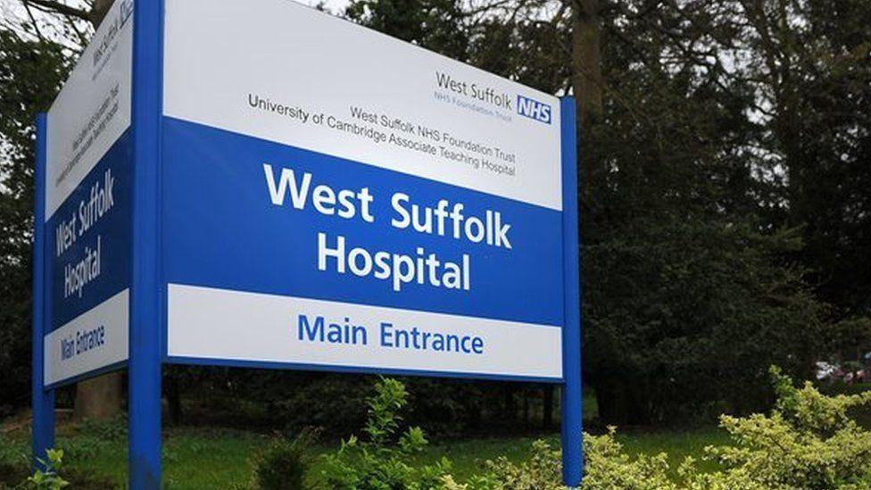 An image of the West Suffolk Hospital main entrance sign. It shows a large blue and white sign in front of a row of trees with bushes surrounding it. 