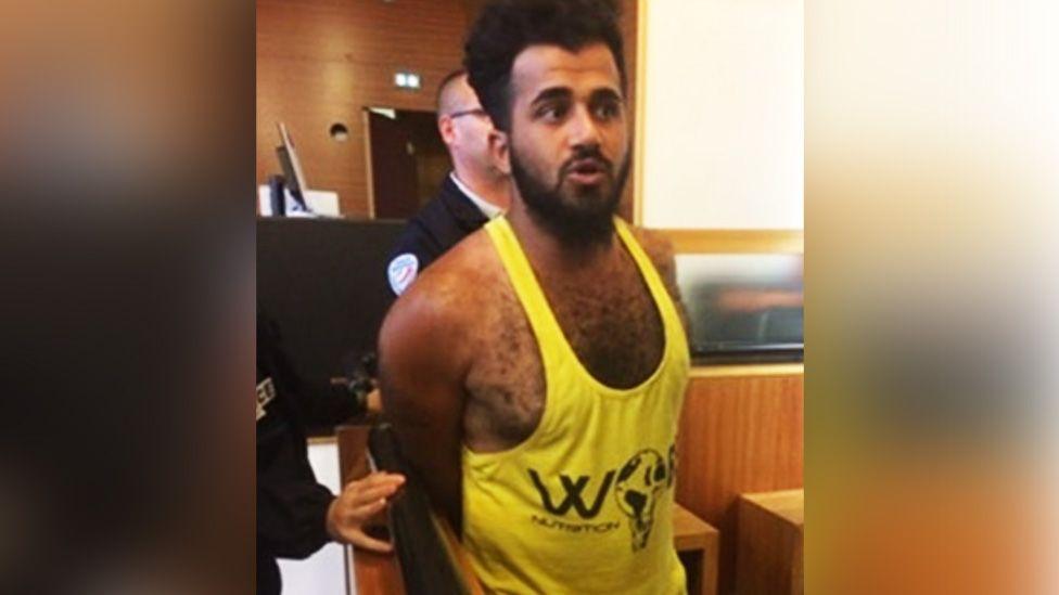 Twana Jamal in a yellow weightlifter's vest with a black graphic stands in an indoor public setting with hands held behind the back, while another individual in dark clothing stands close behind; wood-panelled walls and a counter are visible in the background, and partial text reading “W” appears on the shirt.