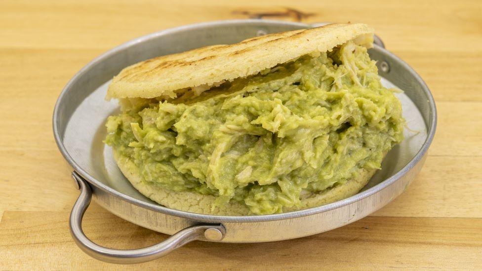 A photo of an arepa, made with yellow corn flour bread, served with a green avocado and chicken mix inside