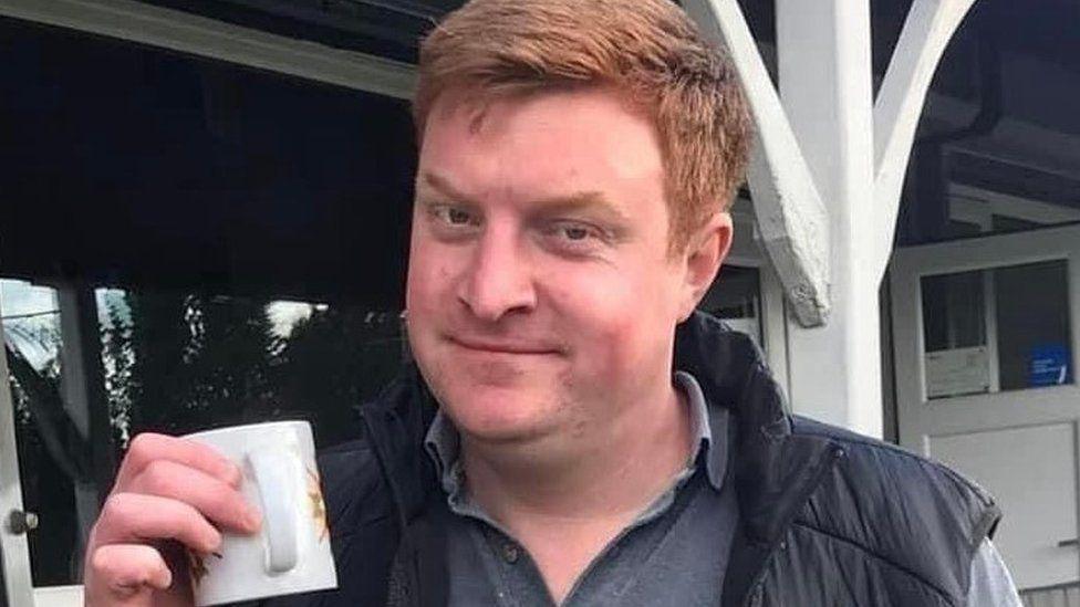 A man with ginger hair, wearing a black jacket and a grey shirt, holds up a mug.