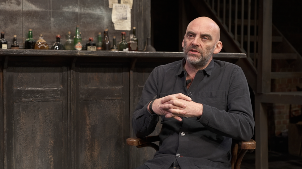 Sean Holmes sitting down on theatre stage. The sets an old western bar