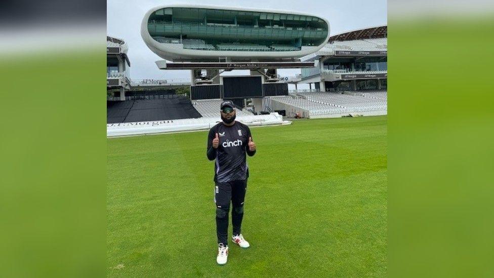 Moshfique Ahmed, a man with a black beard and wearing a black t-shirt and baseball camp and a pair of sunglasses, stands on a cricket pitch giving a thumbs up to the camera.