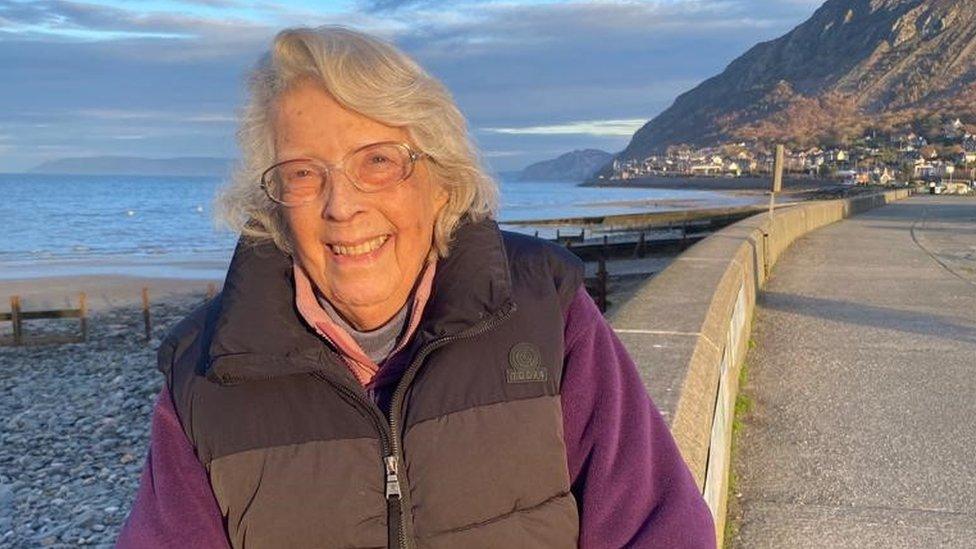 Jo Collie sitting on a wall with a shingle beach behind her