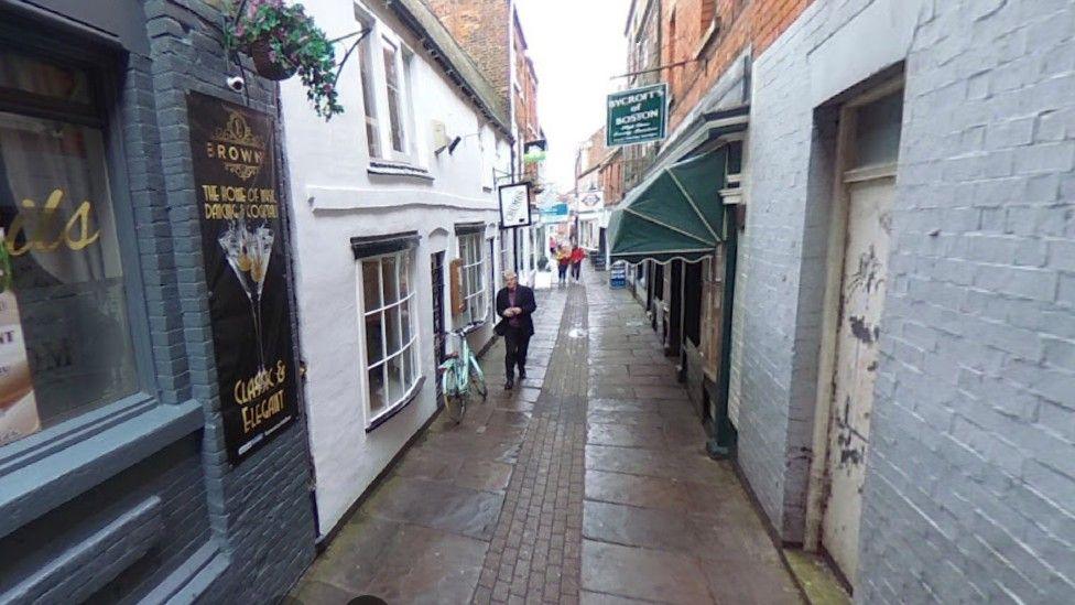 Generic Google shot of a narrow alley with bars and small shops with a brightly coloured bicycle and people in view
