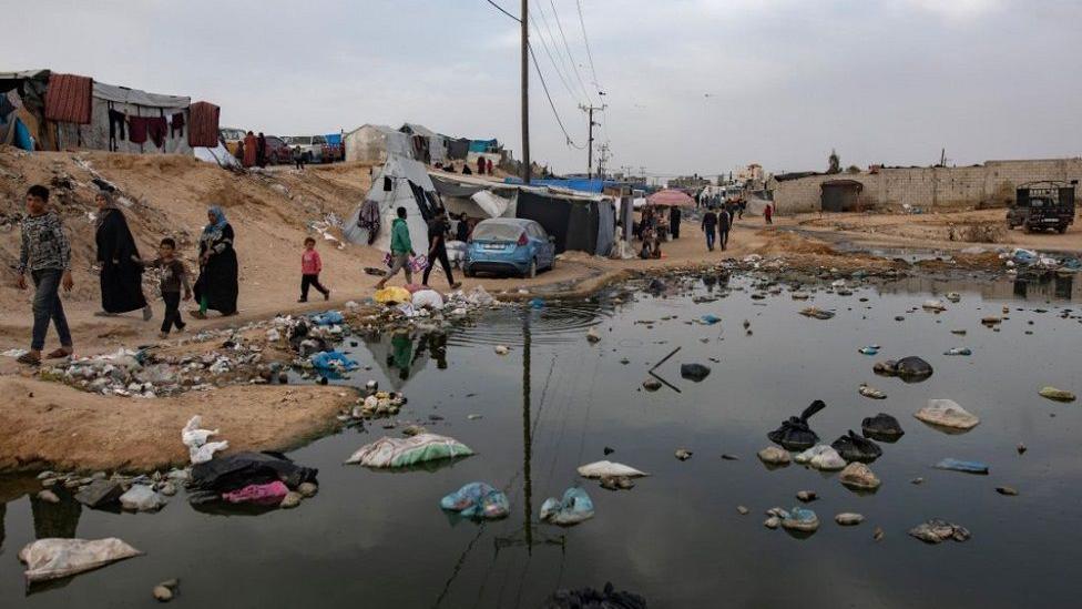 People walk next to a sewage spill and garbage near tents for internally displaced people at a temporary camp in Rafah camp, southern Gaza Strip