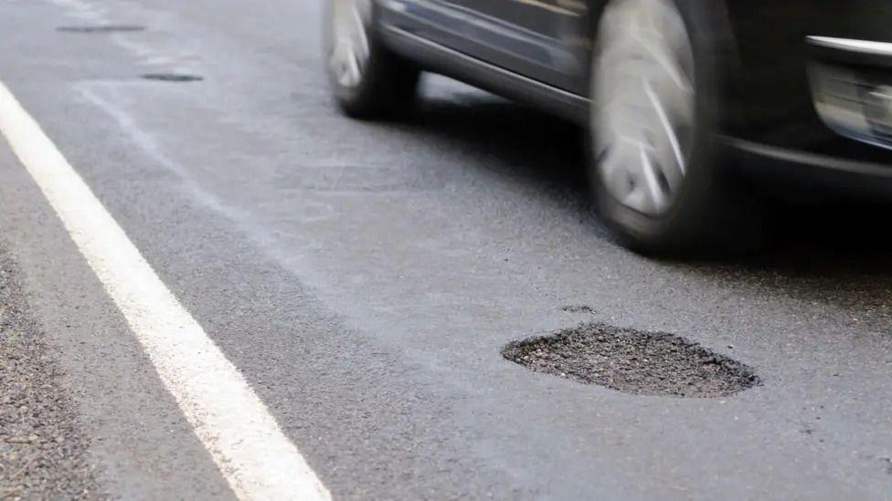 A close up image of a pothole on a road with a car wheels driving close to it